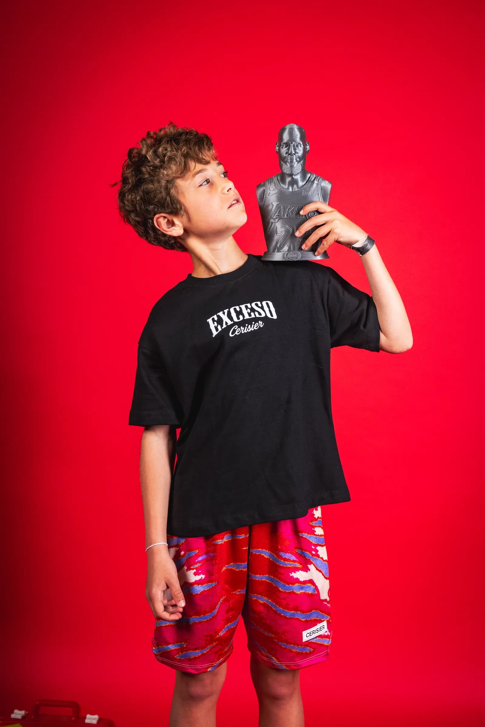 Chico con remera negra y short estampado rojo, sosteniendo un trofeo sobre fondo rojo